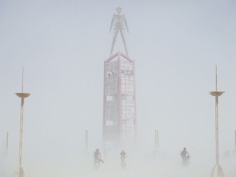 Burning Man towering over burners on bikes in a dust storm.